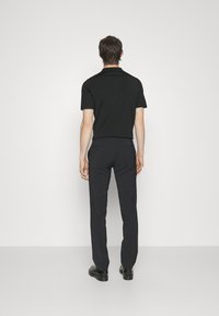 Černé polo tričko s krátkým rukávem spárováno s vypasovanými tmavými kalhotami, z pohledu zezadu. Jemná struktura, obtažený střih a minimální designové detaily.