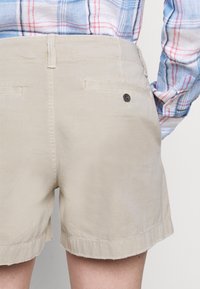 Osoba v béžových šortkách s zadními kapsami a modro-růžovo-bílém kostkované košili s dlouhým rukávem zastrčené dovnitř.