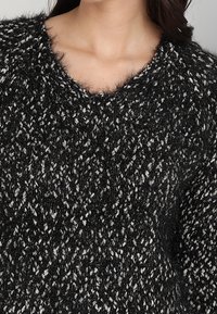 Czarny i biały sweter dzianinowy z dekoltem w serek; puszysta tekstura; luźny splot; kontrastujące włókna tworzą plamisty wzór.