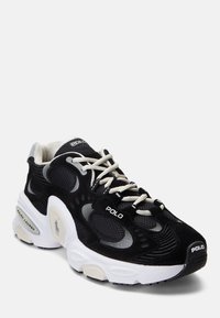 Svart idrottssko med mesh- och mockadetaljer, vit sula, grå accenter och beigefärgade snören. Har en logotyp på sidan och på tungan.