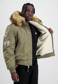 Giacca bomber verde oliva con cappuccio foderato in pelliccia, chiusura con zip e polsini a costine. Presenta toppe ricamate e un morbido interno color crema.