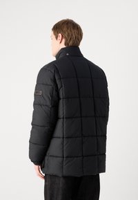 Black puffer jacket with a high collar, square quilted design, and a logo patch on the left sleeve. Material appears textured and insulated.