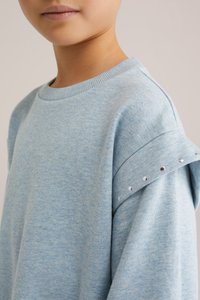 Svijetloplava dukserica s rebrastim ovratnikom i dekorativnim srebrnim zakovicama na ramenu. Mekana tekstura, opušteni kroj i istaknuti šavovi.