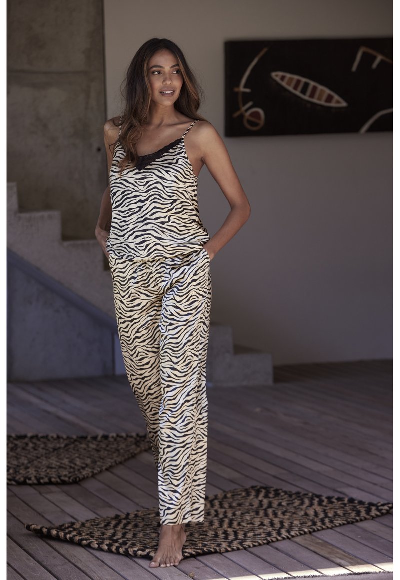 Mouwloze top met zebradessin en zwarte rand, gecombineerd met bijpassende zebradessinpantalon. Zachte, lichte stof. Blootsvoets op een houten vloer.