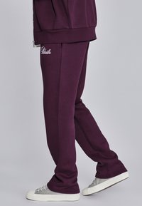 Violeta sporta bikses ar sānu šuvēm un elastīgām piedurknēm, kurām ir balta logo izšūšana uz kreisā augšstilba. Valkātas ar pelēkiem sporta apaviem.