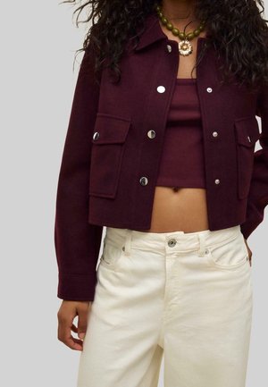 Woman wearing a cropped dark burgundy jacket with silver buttons, matching tank top, white pants, and green beaded necklace with flower pendant.