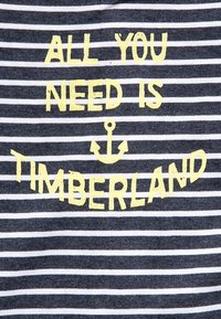 Tričkovina s proužky s žlutým textem "ALL YOU NEED IS TIMBERLAND" s kotvou mezi slovy.