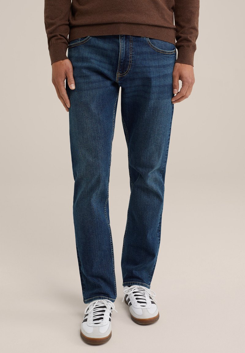 Jeans in denim blu con un design classico, cinque tasche e cuciture gialle a contrasto, abbinati a sneakers bianche.