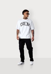 Weißes T-Shirt mit kurzen Ärmeln und schwarzem "ESTEEM"-Grafik, kombiniert mit schwarzen Cargo-Hosen und weißen Sneakern. Lässiger Streetwear-Stil.