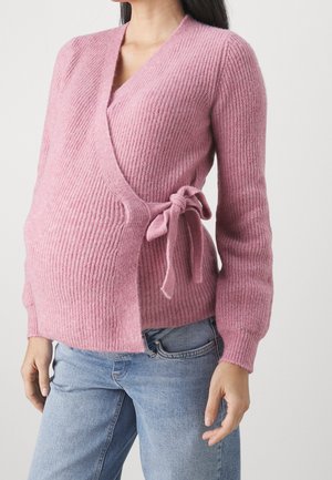 Femme portant un pull en maille rose pâle cache-cœur noué sur le côté, par-dessus un jean bleu clair, montrée du milieu du torse au cou.