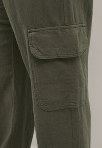 Jambe de pantalon cargo vert olive foncé avec une grande poche rectangulaire à rabat sur le côté, montrant des coutures détaillées et la texture du tissu.