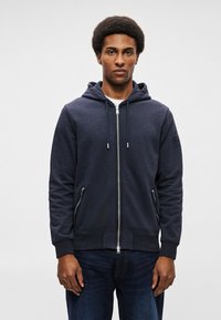 Mornarska zip-up dukserica s kapuljačom na vezanje, bočnim džepovima i rebrastim manžetama. Lagani materijal s glatkom teksturom i minimalnim brendingom.