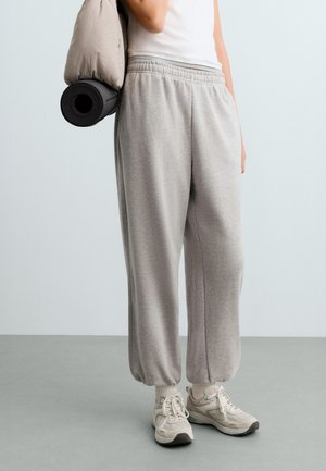 Personne portant un pantalon de survêtement gris et des baskets blanches, tenant une veste beige avec un tapis de yoga noir roulé sur l'épaule.