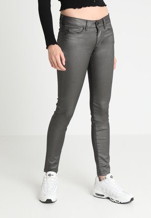 Femme portant un jean slim gris foncé brillant, un top noir à manches longues court, et des baskets blanches Nike Air Max, debout devant un fond blanc.