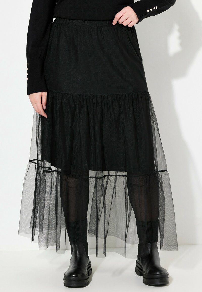 Jupe noire en maille à volants portée avec un top noir, des chaussettes côtelées noires et des bottines chunky en cuir noir sur fond blanc.