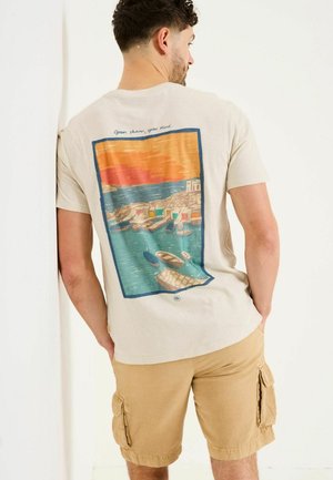Beige Baumwoll-T-Shirt mit buntem Strandmotiv auf der Rückseite, mit Text über dem Bild. Kombiniert mit beigen Cargo-Shorts.