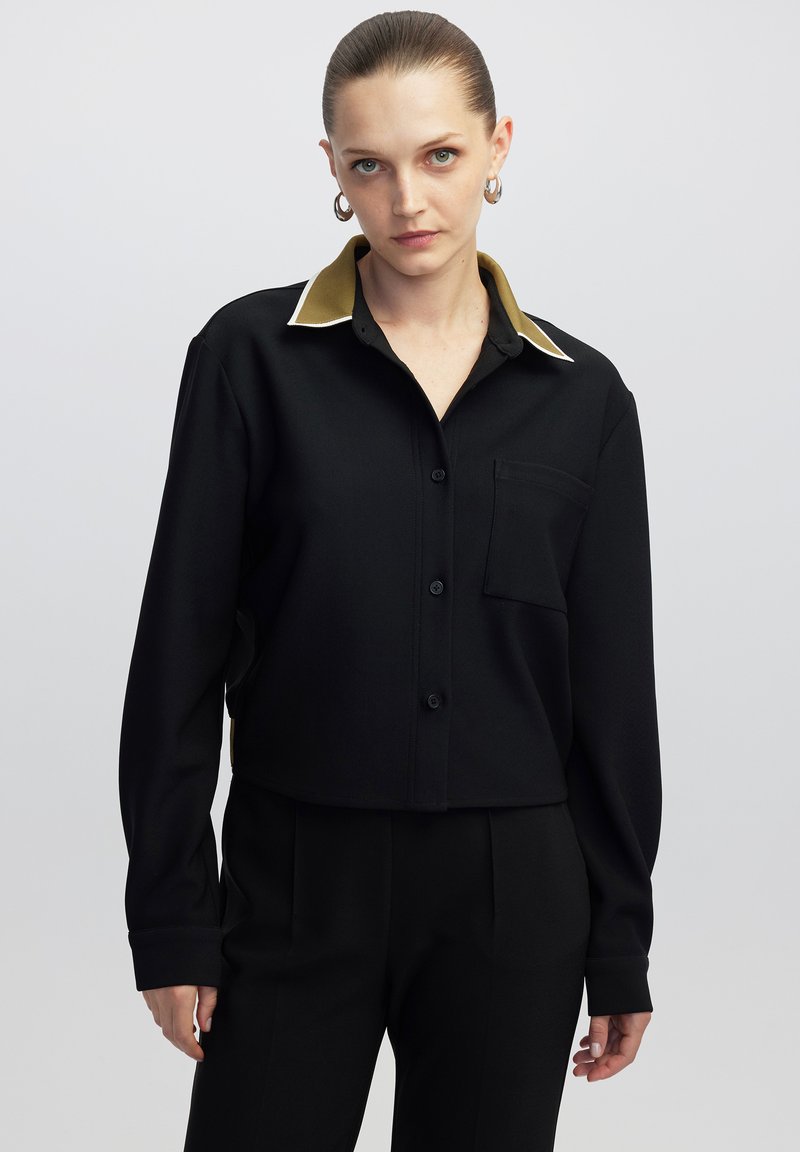 Zwarte cropped shirt met een gouden kraag en witte accenten, met lange mouwen, een zak aan de voorkant en knoopsluiting. Soepele textuur.