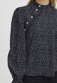 Blusa nera con pois bianchi, dettaglio arricciato e accenti di perle al colletto. Maniche lunghe con polsini arricciati. Tessuto liscio.