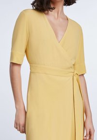 Gul, omslagsklänning med korta ärmar, djup V-ringning och midjeband. Tyget har en slät, lätt strukturerad yta.
