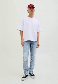 Mlad moški stoji oblečen v rdečo kapo, belo preveliko majico, raztrgane svetlo modre jeans hlače in črne superge pred enobarvno svetlo ozadje.