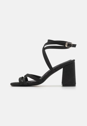 Sandale noire à brides avec un talon bloc, en matériau daim doux, avec des brides croisées et une fermeture à boucle au niveau de la cheville.