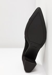 Chaussure à talon haut en daim noir à bout pointu, placée semelle vers le haut contre une surface et un mur blancs.
