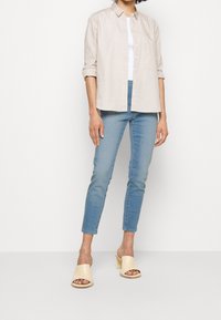 Camisa botón de un color beige claro con mangas remangadas, llevada sobre una camiseta blanca sin mangas. Jeans ajustados de color azul y sandalias de tacón bloque abierto en color beige.