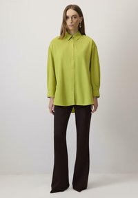 Camicia oversize verde lime con bottoni e colletto, abbinata a pantaloni neri a vita alta svasati, creando una combinazione di colori contrastante.