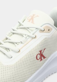 Adidaș Calvin Klein cu un partea superioară din plasă bej, talpă albă, logo roșu, accente crem, șireturi plate și detalii texturate pe suprafață.