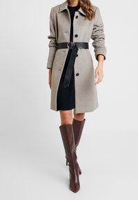 Abrigo de lana gris con cuello de muesca, cinturón de cuero negro, vestido negro debajo y botas de tacón alto marrones hasta la rodilla.