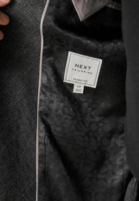 Interior de una chaqueta negra ajustada con forro de patrón geométrico. La etiqueta dice "NEXT TAILORING SKINNY FIT" con detalles sobre tamaño y origen visibles.