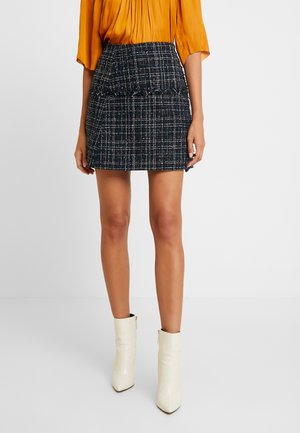 Femme portant une mini-jupe à carreaux noir et blanc, un chemisier jaune moutarde et des bottines blanches, debout devant un fond uni.