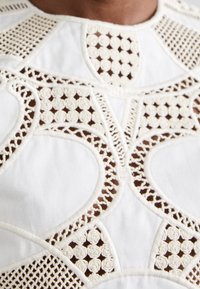 Haut en tissu blanc présentant des découpes en dentelle complexes et des motifs brodés, avec des designs circulaires et courbés et une finition texturée.