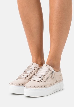 Beżowe sneakersy na platformie z satynowymi sznurowadłami, metalicznymi ćwiekami wokół podeszwy oraz suwakiem, noszone na boso na białym tle.