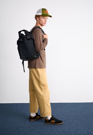 Jeune homme en pantalon beige, chemise marron à manches longues, casquette verte et blanche, chaussures noires et marron, portant un sac à dos noir sur une épaule.