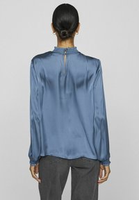 Blusa di seta in blu polveroso con maniche lunghe, scollatura arricciata e dettaglio a chiave sul retro. Il modello la indossa con jeans grigi e testurizzati.