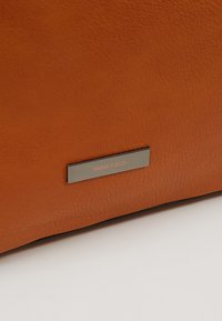 Ruskea nahkalaukku, jossa on sileä pinta, hopeinen metallilaatta, jossa lukee "ANNA FIELD", ja yksinkertainen, jäsennelty muotoilu.