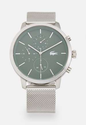 Montre en métal argenté avec un cadran vert, comportant deux sous-cadrans et des index horaires blancs. Elle possède un bracelet en maille et le logo Lacoste à la position de 6 heures.