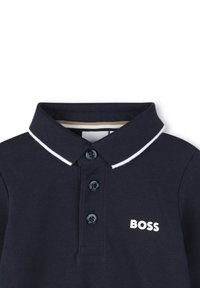 Polo-shirt bleu navy foncé avec un tissu texturé, bord col blanc contrasté, trois boutons navy et logo "BOSS" en blanc sur la poitrine.