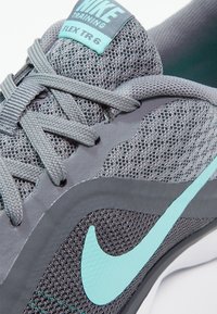 Zapatillas de entrenamiento Nike grises con parte superior de malla, cordones grises, una etiqueta azul verdoso y un detalle de swoosh azul verdoso sobre una suela flexible.