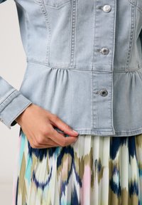 Hellblaue Jeansjacke mit einem Peplum-Design, silbernen Beschlägen und einer Hand, die den Saum anpasst, kombiniert mit einem bunten, gemusterten Rock.