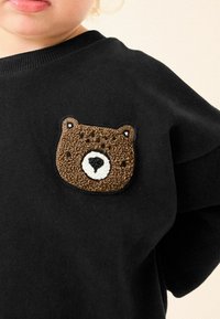 Sweatshirt noir avec une broderie en forme d'ours brun sur la poitrine gauche, présentant des détails de fourrure texturée et un museau blanc.