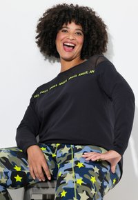Zwarte lange mouw top met mesh-accenten en neon groene tekst; gecombineerd met blauwe camouflagebroek met gele sterren en groene stiksels.