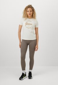 Kremhvitt t-skjorte med brun "Guess" logo, kombinert med teksturerte brune leggings og svarte sneakers, med grønne detaljer og hvite sokker.