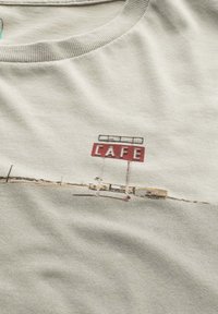 Lichtgrijs katoenen T-shirt met een geborduurd ontwerp van een rood "CAFE"-bord en een eenvoudig landschap, met subtiele textuur en zachte stof.