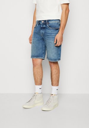 Homme portant un short en jean bleu mi-long, des chaussettes Nike blanches mi-mollet et des baskets Nike montantes de couleur claire, debout devant un fond uni.
