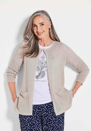Cardigan tricoté en beige clair avec un tissage ouvert, doté de deux poches avant et de manches 3/4, superposé sur un t-shirt graphique blanc et un pantalon à pois.