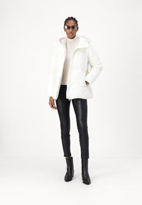 Hvid pufferjakke med hætte, med store frontlommer og lynlås. Bæres over en beige sweater med sorte strukturerede skinny jeans og støvler.