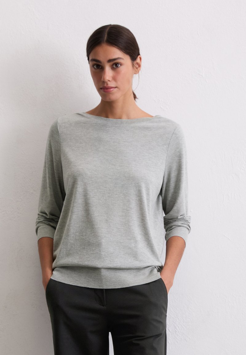 Hellgrauer, langärmliger Pullover mit weitem Ausschnitt und weicher Textur, kombiniert mit dunklen Hosen, die einen lässigen Schnitt und hochgekrempelte Ärmel aufweisen.