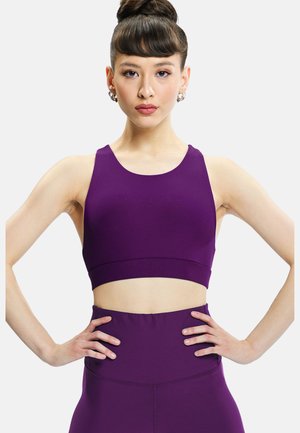 Woman with hair in a bun wearing matching purple crop top and high-waisted leggings, standing with hands on hips against white background.